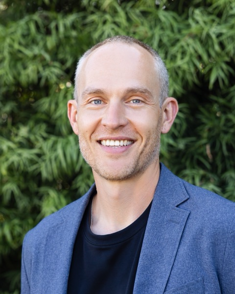Blake Lawson smiling, in a blue blazer and black T-shirt, in front of green foliage